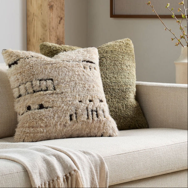 Two textured throw pillows on a light beige sofa in a styled living room setting. The front pillow is cream with black geometric woven patterns, and the back pillow is olive green.