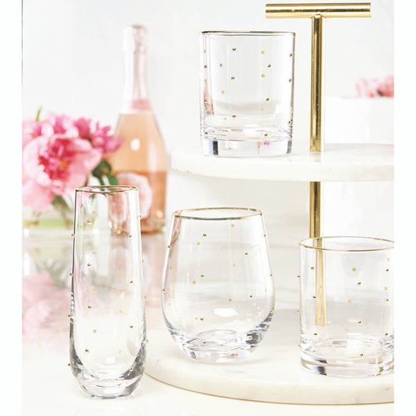 Set of clear glasses with gold dots on a decorative stand with flowers and a bottle in the background.