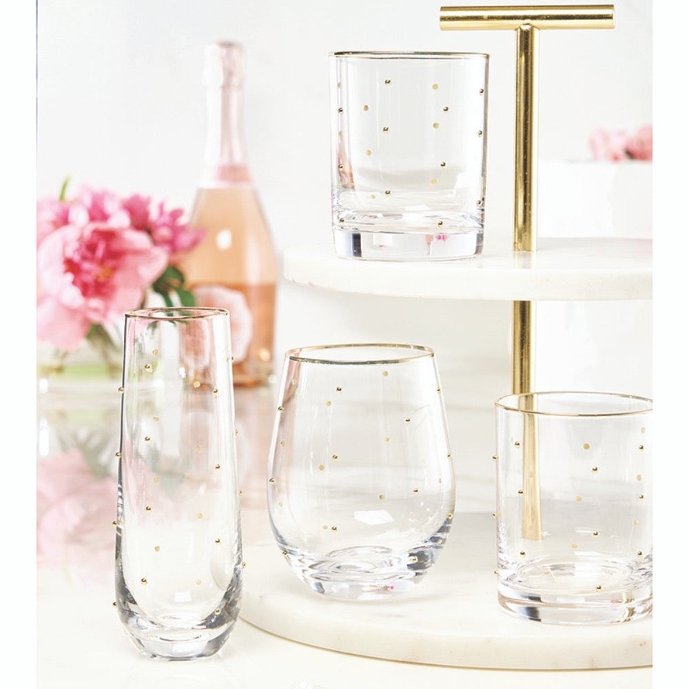 Set of clear glasses with gold dots on a decorative stand with flowers and a bottle in the background.
