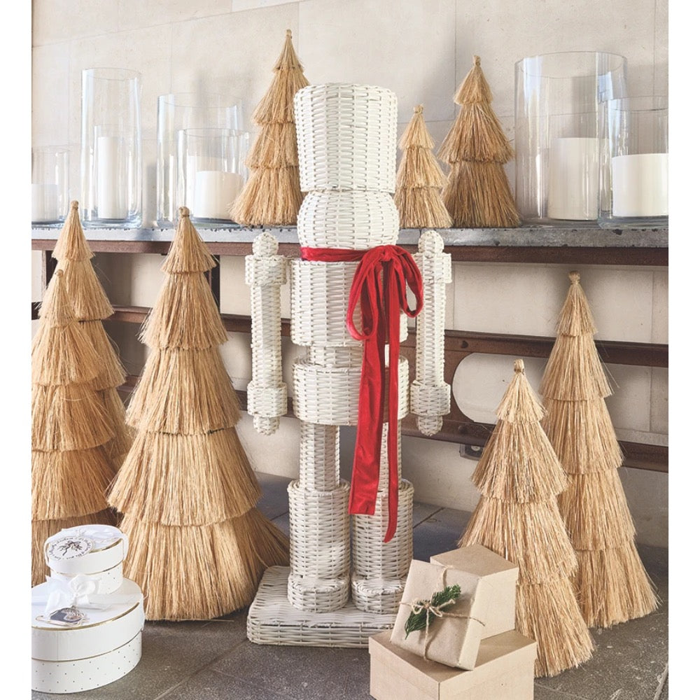 A white woven nutcracker decoration tied with a red ribbon, displayed among natural straw Christmas trees and wrapped gift boxes on a shelf.