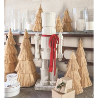 A white woven nutcracker decoration tied with a red ribbon, displayed among natural straw Christmas trees and wrapped gift boxes on a shelf.