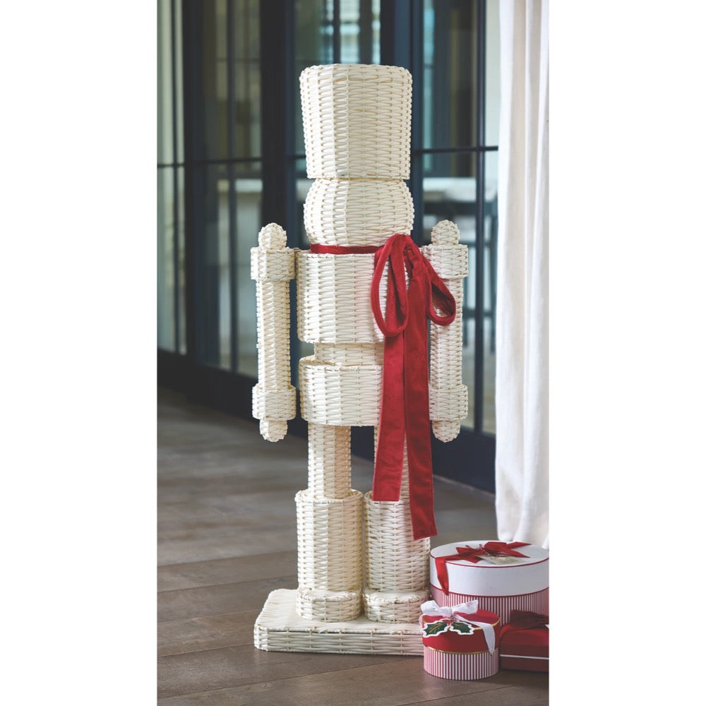 A tall, rustic nutcracker-shaped decorative figure made from woven natural wood or wicker, with a bright red ribbon tied around the waist and small gift boxes at its base, set indoors near a window and curtain.