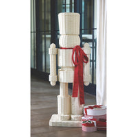 A tall, rustic nutcracker-shaped decorative figure made from woven natural wood or wicker, with a bright red ribbon tied around the waist and small gift boxes at its base, set indoors near a window and curtain.