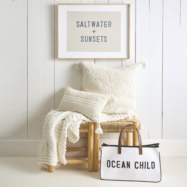 Chair with pillows and a blanket next to a bag with 'Ocean Child' text, against a white wall with a framed picture.