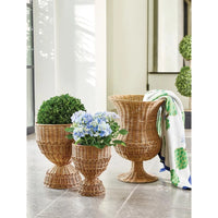 Three woven wicker planter pots in graduated sizes on a tiled floor, with two containing green boxwood plants and one holding blue and white flowers. A white throw blanket with green and blue botanical print is draped over the largest pot.