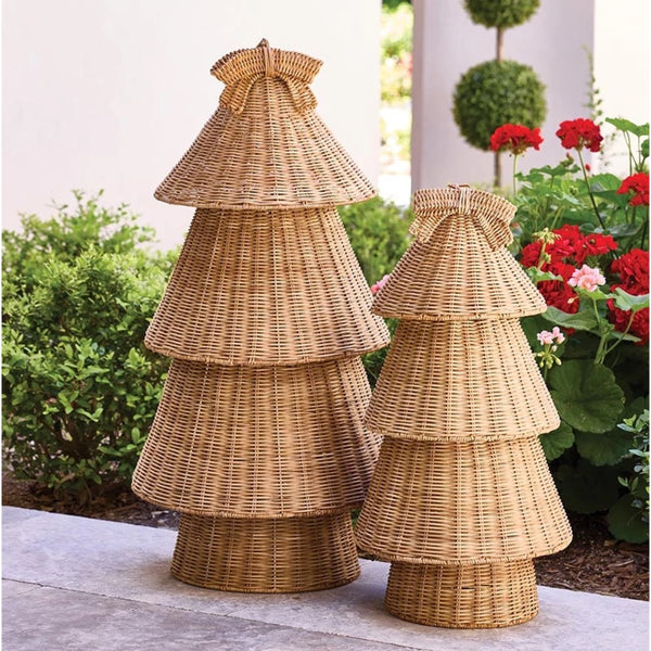 Two decorative tiered wicker tree ornaments in natural tan tones, displayed outdoors on a stone surface with green shrubs and red flowers in the background.
