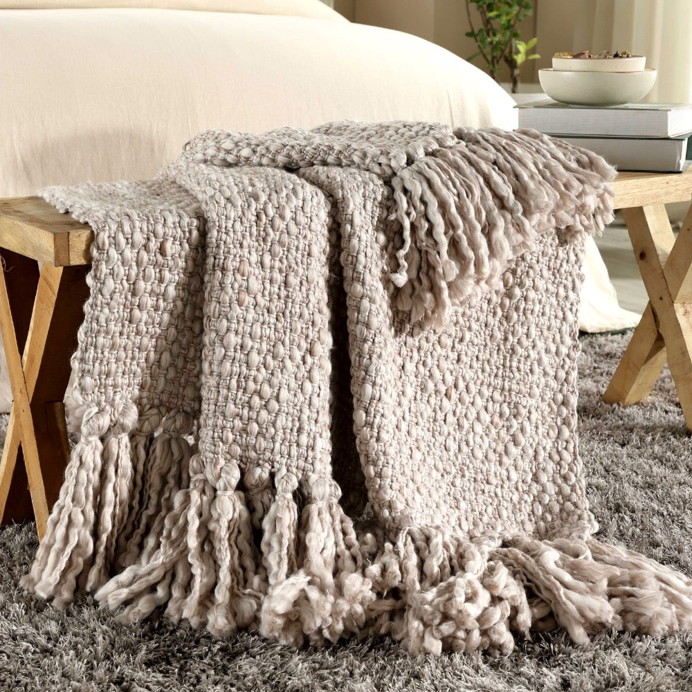 India's Heritage Montana Natural Wool Throw with fringe draped over a wooden bench next to a bed and shaggy gray rug.