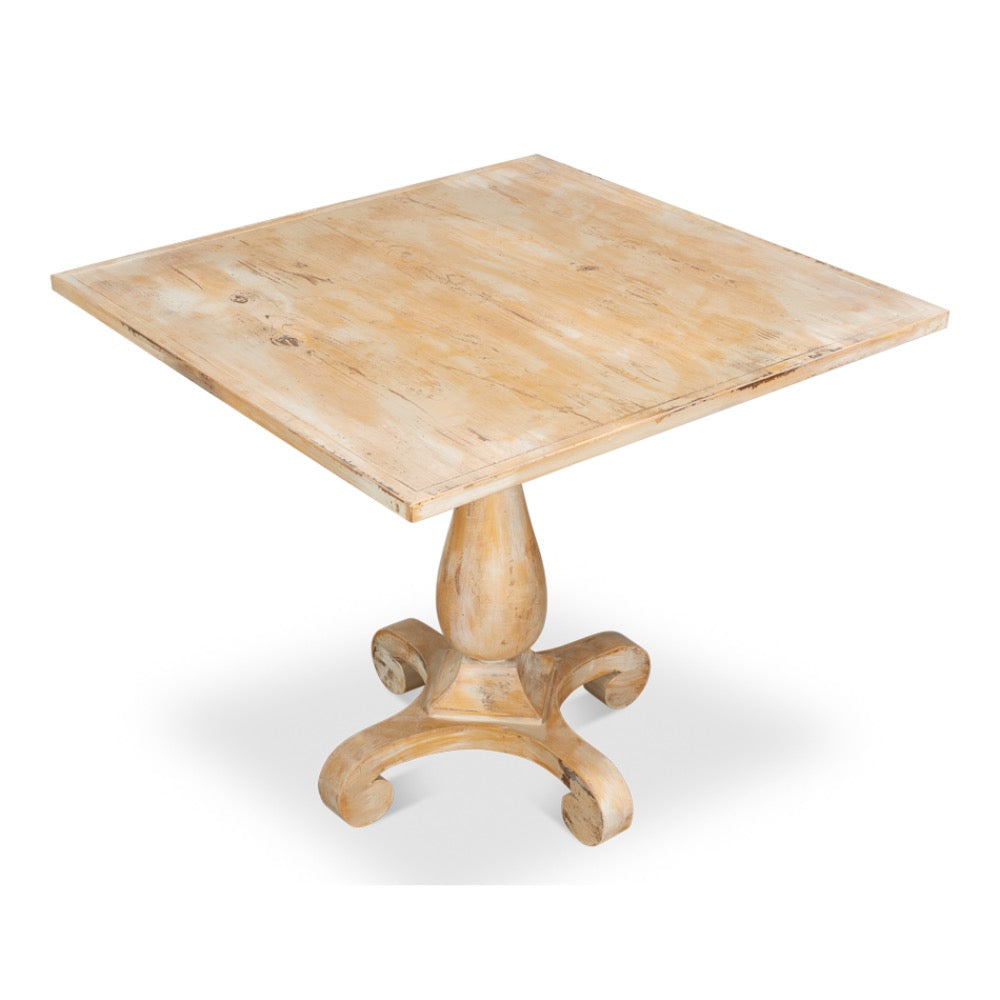 A square wooden pedestal table with a distressed light beige finish, featuring a turned central base and four curved legs, shown on a white background.