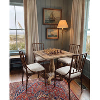 A rustic wooden dining table with four matching spindle-back chairs in a coastal-style room, set on a patterned rug with windows and framed sailboat paintings in the background.