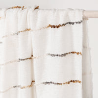 A close-up of a white textured throw blanket draped over a light wooden rod, featuring horizontal rows of small metallic beads in gold, bronze, and silver.
