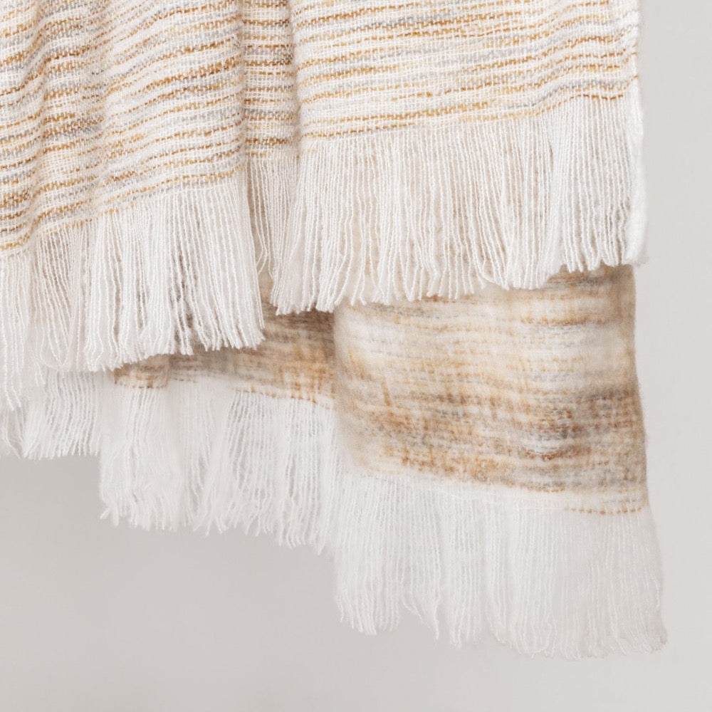 A close-up of a woven throw blanket with white fringe, featuring horizontal stripes in cream, beige, and light brown.