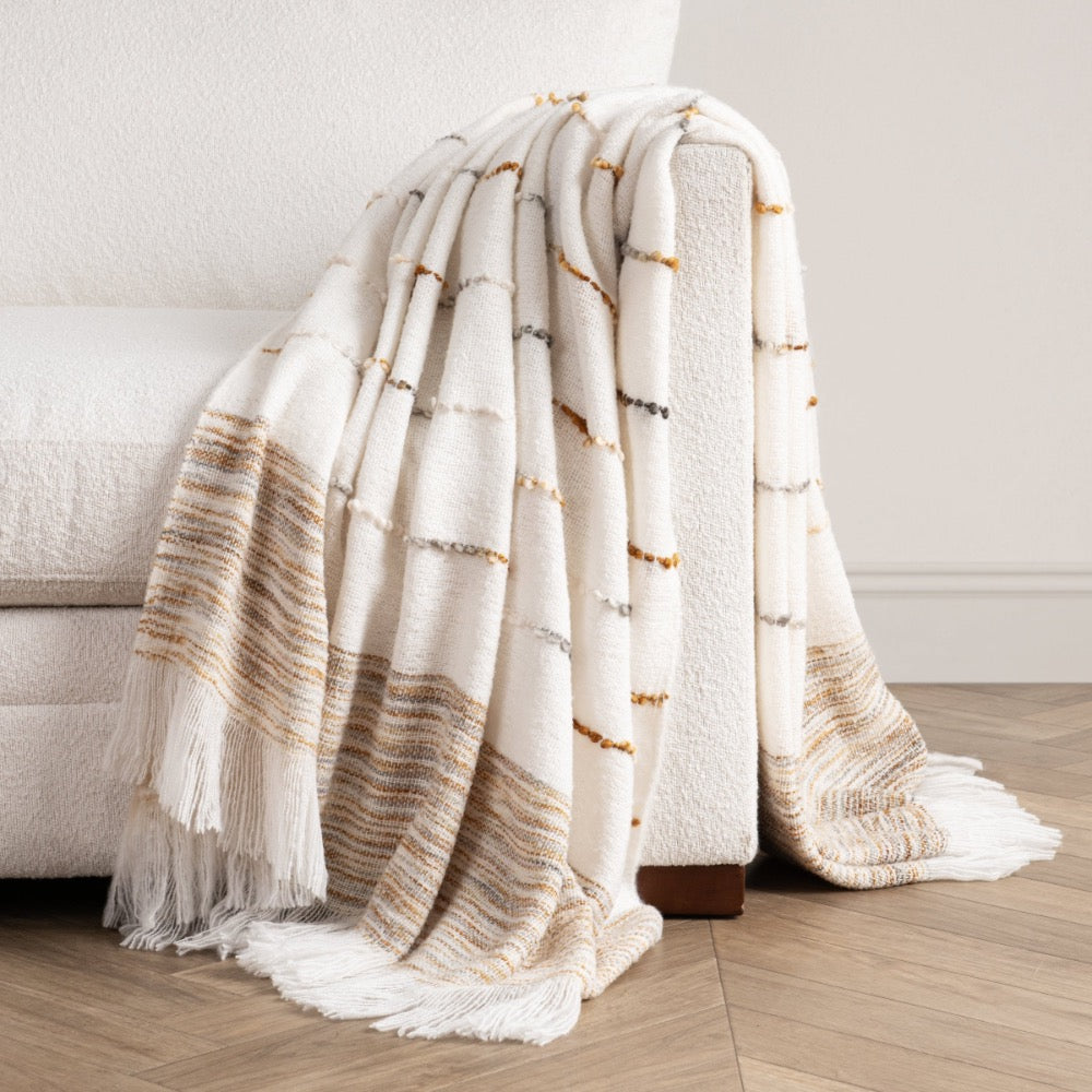 A textured cream throw blanket with fringe and horizontal striped patterns in beige, brown, and gray, draped over the arm of a white sofa on a light wood floor.
