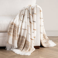 A textured cream throw blanket with fringe and horizontal striped patterns in beige, brown, and gray, draped over the arm of a white sofa on a light wood floor.