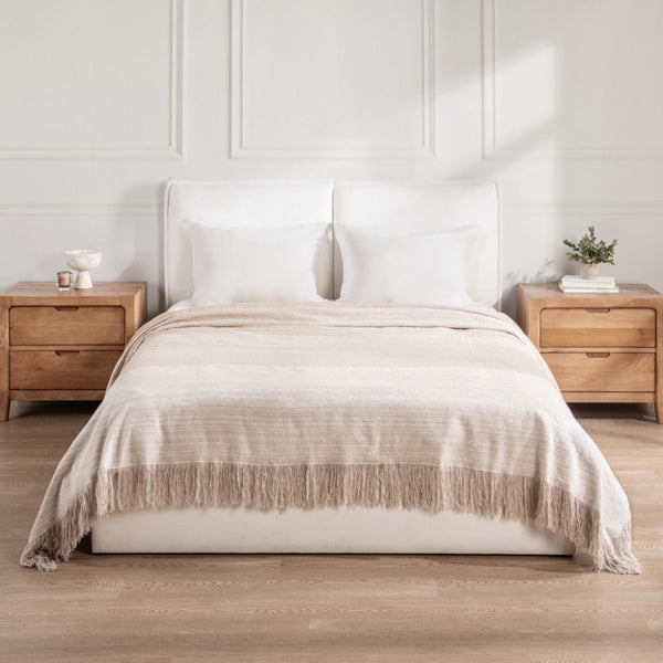 Image of a bed made with beige blanket with fringe and two wooden nightstands on each side on a white wall.