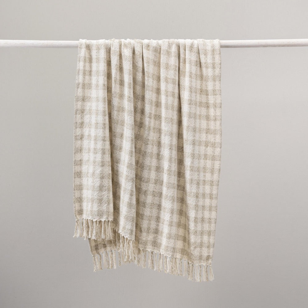 A cream and beige gingham check throw blanket with fringed edges hanging from a white horizontal rod against a plain light gray background.