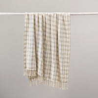 A cream and beige gingham check throw blanket with fringed edges hanging from a white horizontal rod against a plain light gray background.