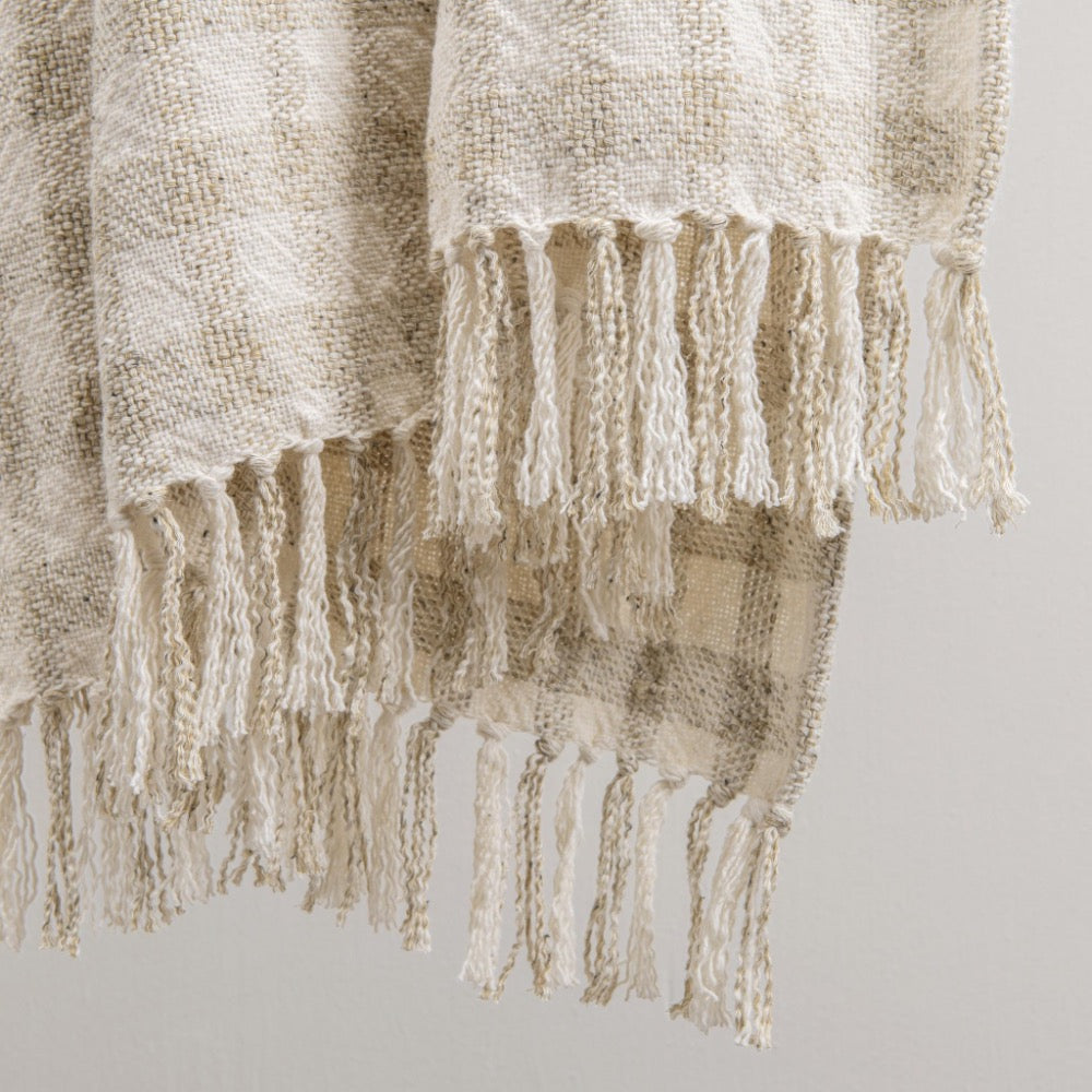 A close-up of a woven throw blanket with fringed edges, shown draped against a plain light gray background. The blanket has a textured, natural-looking weave in warm off-white and beige tones.