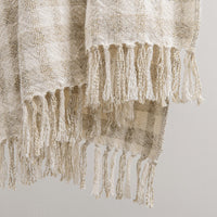 A close-up of a woven throw blanket with fringed edges, shown draped against a plain light gray background. The blanket has a textured, natural-looking weave in warm off-white and beige tones.