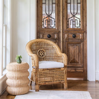 Wicker armchair with a cushion next to a textured side table in a room with wooden doors.