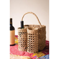 A natural woven straw wine carrier with a single arched handle, holding two wine bottles, set on a colorful quilted blanket with a wine bottle blurred in the background.