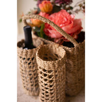 Woven seagrass basket with wine bottle holders with a handle, shown holding wine bottles and flowers in a softly lit setting.