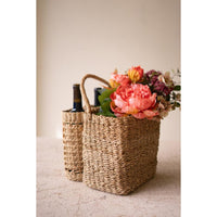 Woven natural-fiber basket on a light stone surface against a warm beige wall. The front basket is filled with a peach-pink floral arrangement and greenery, and a second basket stands behind it holding dark bottles.