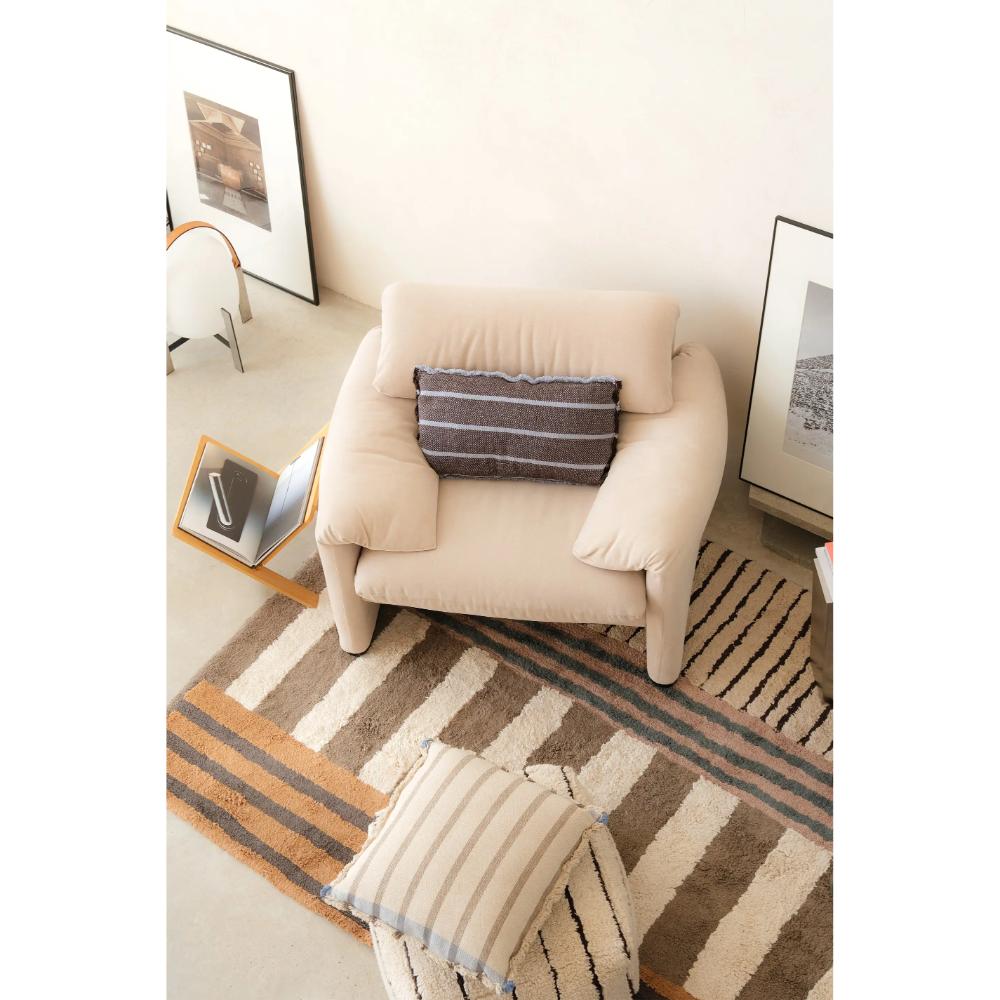 A beige upholstered armchair in a styled room setting, featuring a dark gray striped throw pillow. The chair is placed on a brown and beige striped area rug, with a matching striped throw pillow in the foreground and framed wall art on the walls.