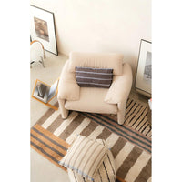 A beige upholstered armchair in a styled room setting, featuring a dark gray striped throw pillow. The chair is placed on a brown and beige striped area rug, with a matching striped throw pillow in the foreground and framed wall art on the walls.