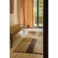 A rectangular area rug with a striped pattern in beige, brown, and muted pink, styled in a warm, sunlit room with a bench and curtains.