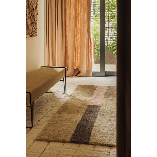 A rectangular area rug with a striped pattern in beige, brown, and muted pink, styled in a warm, sunlit room with a bench and curtains.