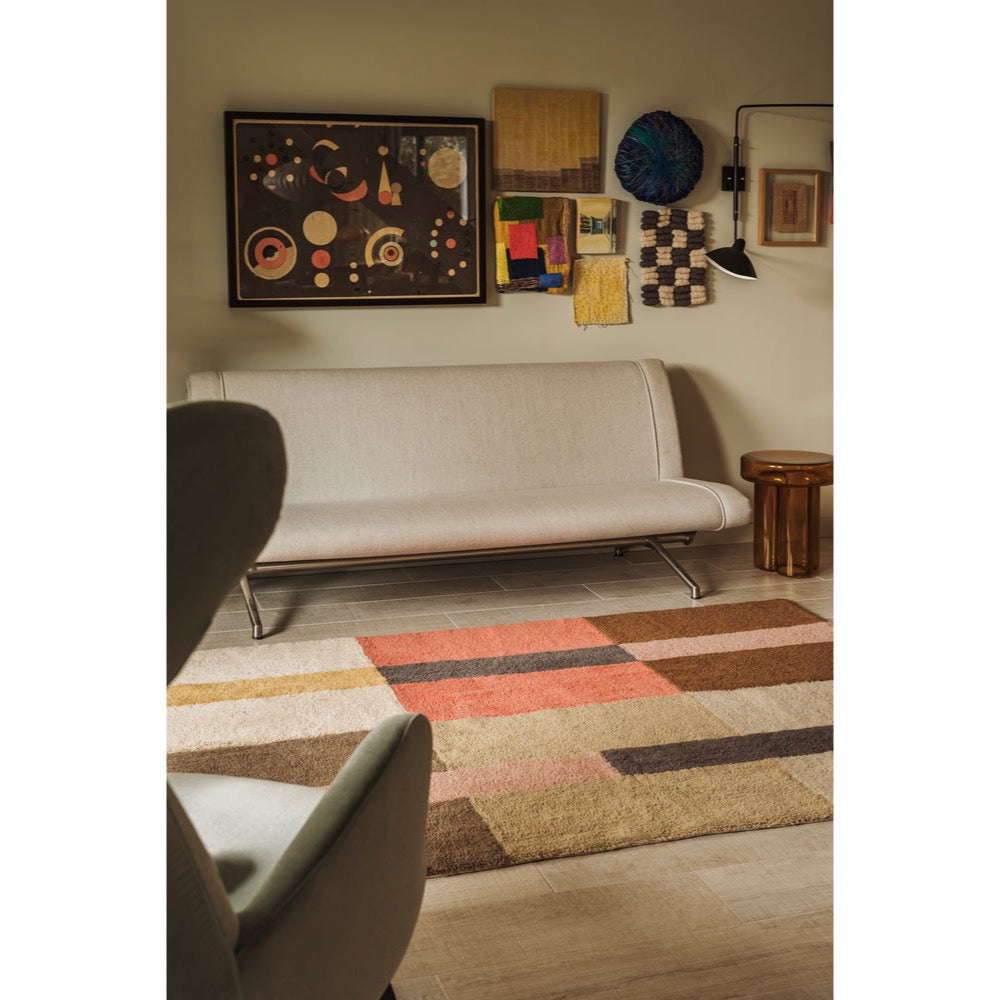 A modern living room scene featuring a low beige upholstered sofa, a colorful geometric area rug with blocks of rust, brown, beige, and gray, and wall art above the couch.