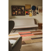 A modern living room scene featuring a low beige upholstered sofa, a colorful geometric area rug with blocks of rust, brown, beige, and gray, and wall art above the couch.