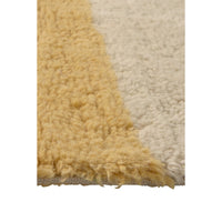 Close-up of a shaggy rug corner showing two color blocks: a warm mustard yellow section and an off-white/cream section, with a fringed edge visible against a white background.