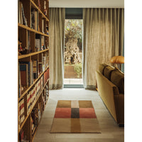 A rectangular area rug with a patchwork design in beige, brown, rust red, and black lies on a light wood floor in a modern living room setting with a bookshelf, sofa, and window.