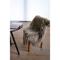 A fluffy sheepskin-style chair cover in a taupe-brown color draped over a wooden chair in a minimalist room with light wood floors and a black side table.