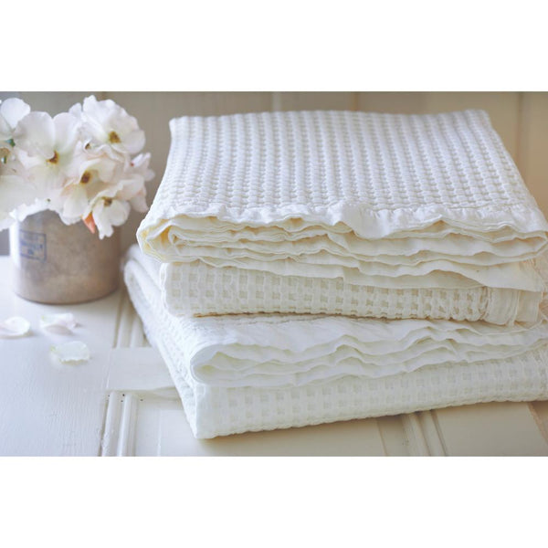 Folded white towels stacked on a table beside a vase of white flowers exude elegance and freshness, much like the Taylor Linens Waffle Weave Blanket made from breathable 100% cotton, inviting calm and comfort into the space.