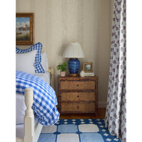 A styled bedroom scene featuring a bed with a blue and white gingham comforter and matching pillow shams, a rustic three-drawer nightstand with a blue ceramic lamp, patterned wallpaper, and a matching blue and white patterned rug with a floral curtain.