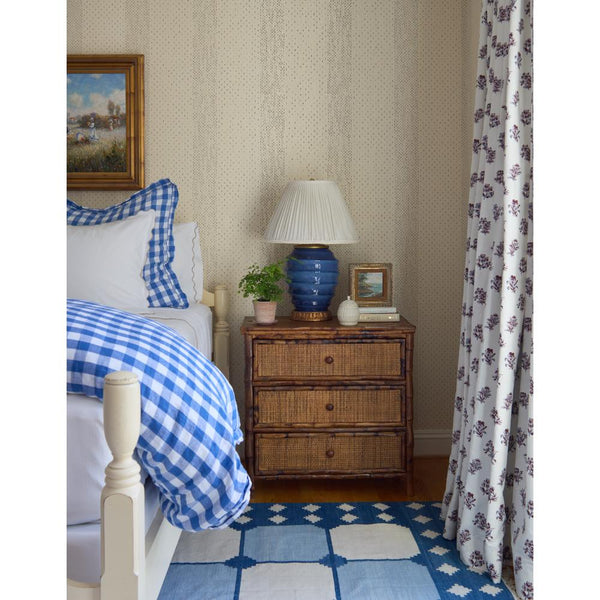 A styled bedroom scene featuring a bed with a blue and white gingham comforter and matching pillow shams, a rustic three-drawer nightstand with a blue ceramic lamp, patterned wallpaper, and a matching blue and white patterned rug with a floral curtain.