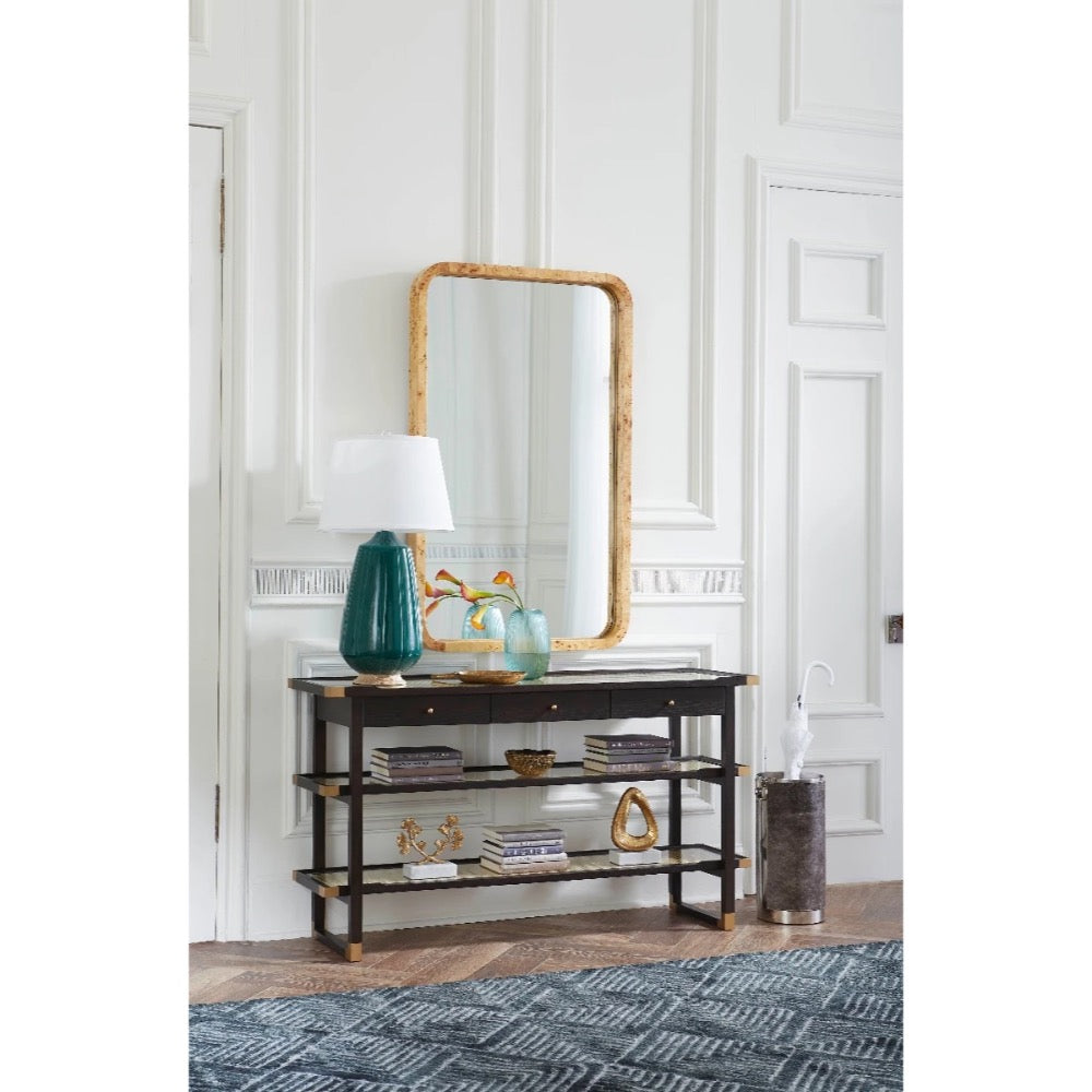 A dark wood console table with three open shelves, styled with a teal lamp, books, and a gold-framed mirror in a white paneled room.