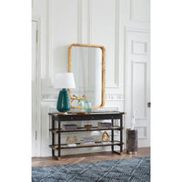 A dark wood console table with three open shelves, styled with a teal lamp, books, and a gold-framed mirror in a white paneled room.