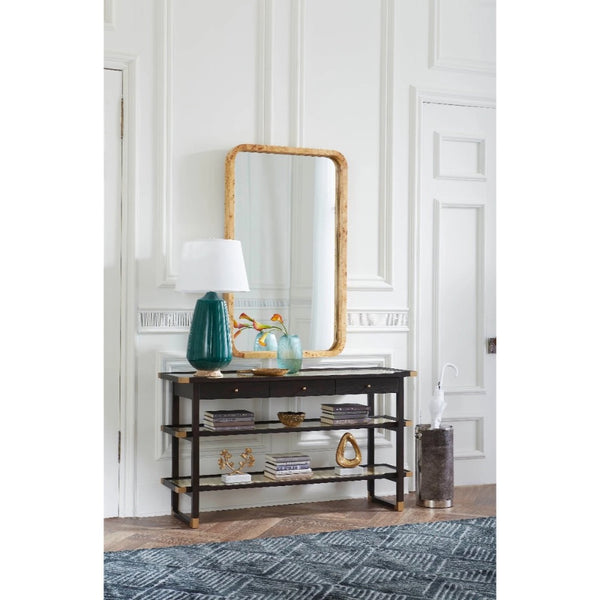 A dark wood console table with three open shelves, styled with a teal lamp, books, and a gold-framed mirror in a white paneled room.