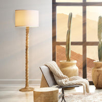 A tall floor lamp with a twisted wooden base and a warm light white fabric shade stands beside a chair in a styled room. A large window shows a desert landscape outside, with potted cacti and a throw blanket adding to the scene.
