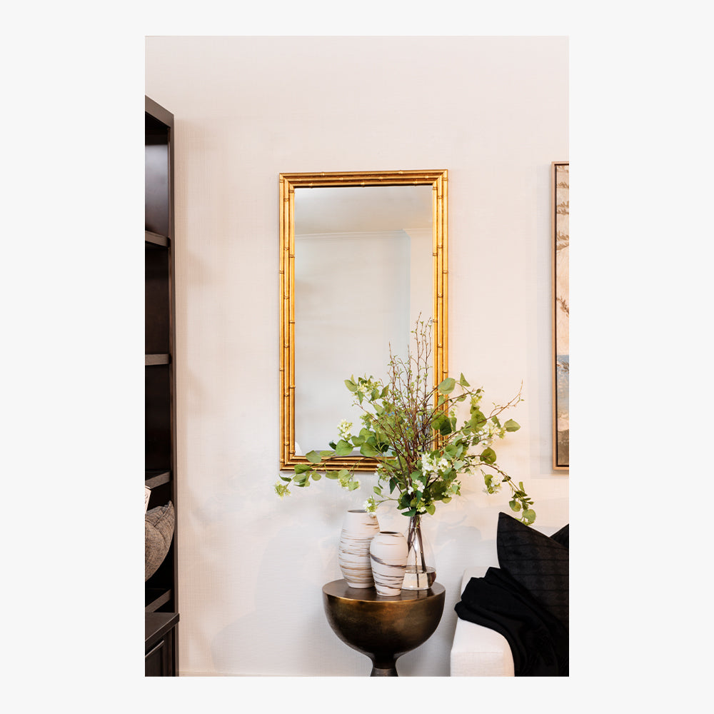 Decorative interior scene with a gold-framed mirror, vase with greenery, and black chair.