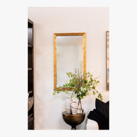 Decorative interior scene with a gold-framed mirror, vase with greenery, and black chair.