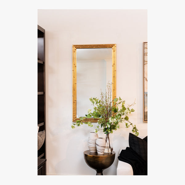 Decorative interior scene with a gold-framed mirror, vase with greenery, and black chair.