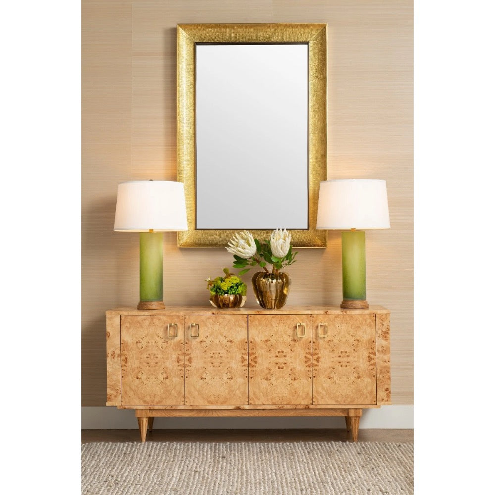 A mid-century modern sideboard cabinet with burl wood grain, flanked by two green table lamps with white drum shades, beneath a large gold-framed mirror on a beige textured wall.