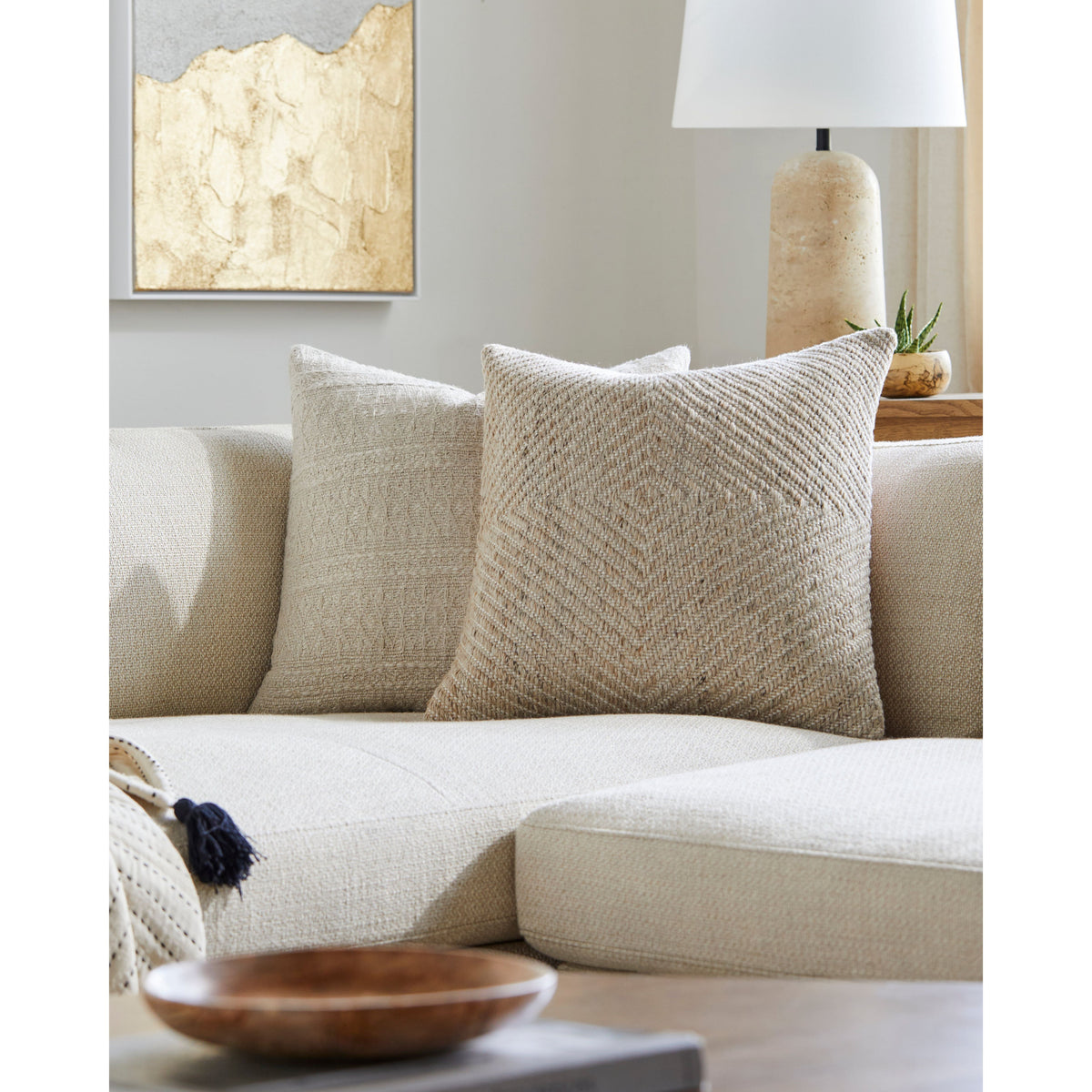 Inviting living room featuring a beige sectional sofa, Surya's Indoor/Outdoor Cairn Woven Pillow crafted from fade-resistant material for added texture, complemented by a wooden bowl and a lamp on the side table.