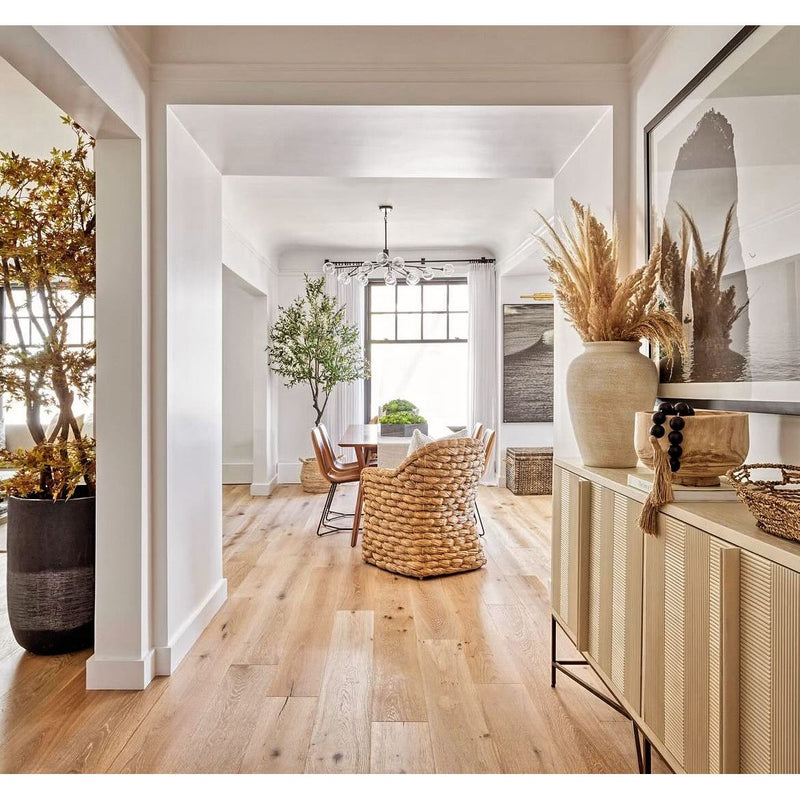 Bright dining room featuring a wooden floor and a modern table, complemented by the Phillips Scott Havana Seagrass Woven Chairs that add a coastal vibe. The space is accented with plants and large vases, while a large window allows natural light to flood in.