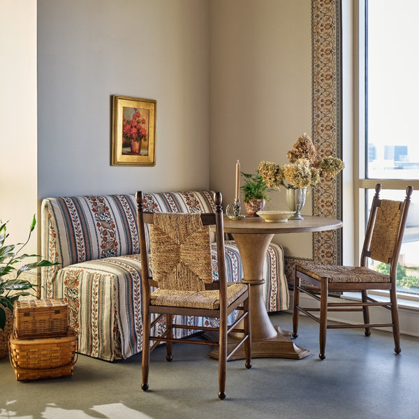 A styled dining nook featuring a round wooden pedestal table, two wooden chairs with woven cane seats, and a matching striped upholstered banquette against a neutral gray wall.