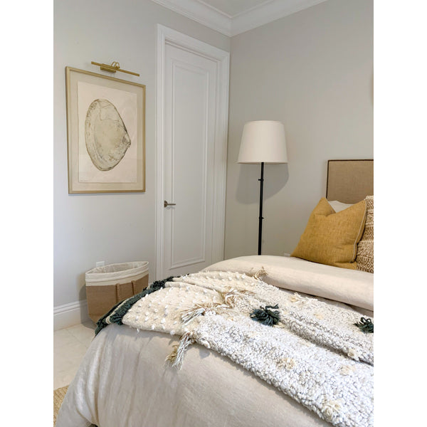 A cozy bedroom features a neatly made bed adorned with decorative pillows and a textured throw. A floor lamp casts light on the Barloga Studios' Atlantic Clam #5 Photography Art, which showcases ocean life motifs and adds a touch of coastal charm to the neutral palette.